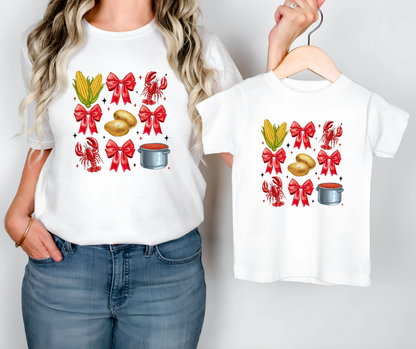 Person holding a white t-shirt with festive designs, including corn, bows, and pots, on a plain background.